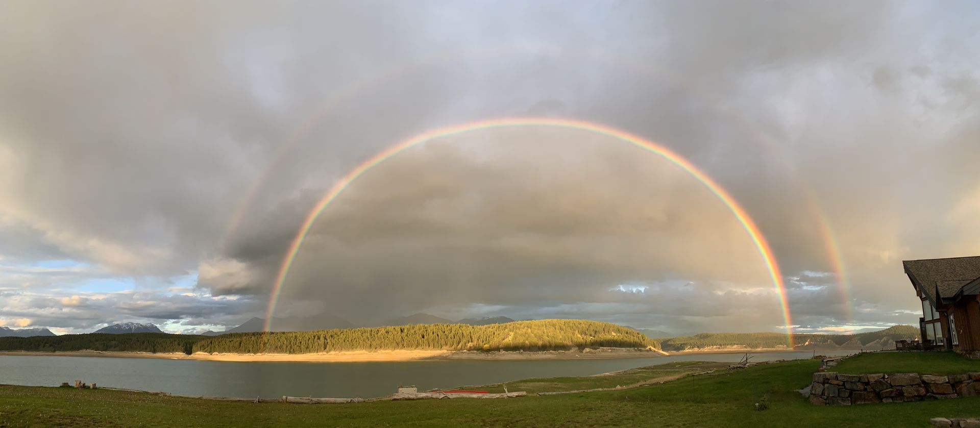 A double rainbow arcing over Lake Koocanusa and the surrounding forest, seen from the village shoreline.